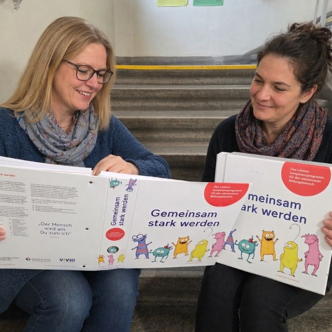Zwei Mitarbeiterinnen von Gemeinsam stark werden sitzen nebeneinander auf einer Treppe, halten die Programmordner für den Elementarbereich in den Händen und blättern lächelnd darin