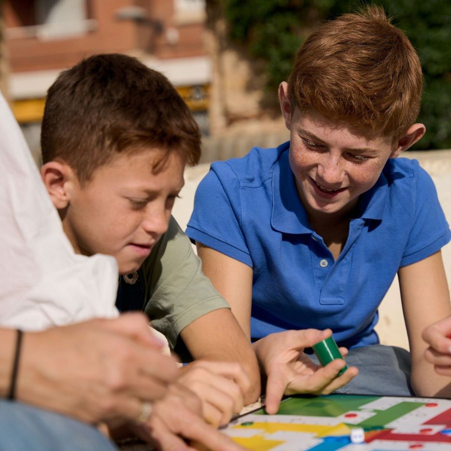 Zwei Buben spielen im Freien gemeinsam mit einer erwachsenen Person ein buntes Brettspiel, einer hält lächelnd eine grüne Spielfigur in der Hand